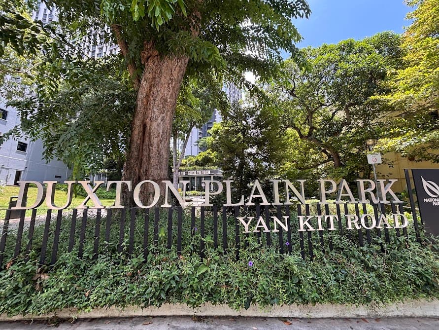 Metal sign lettering reading "Duxton Plain Park Yan Kit Road" set against a black fence, trees, and greenery.