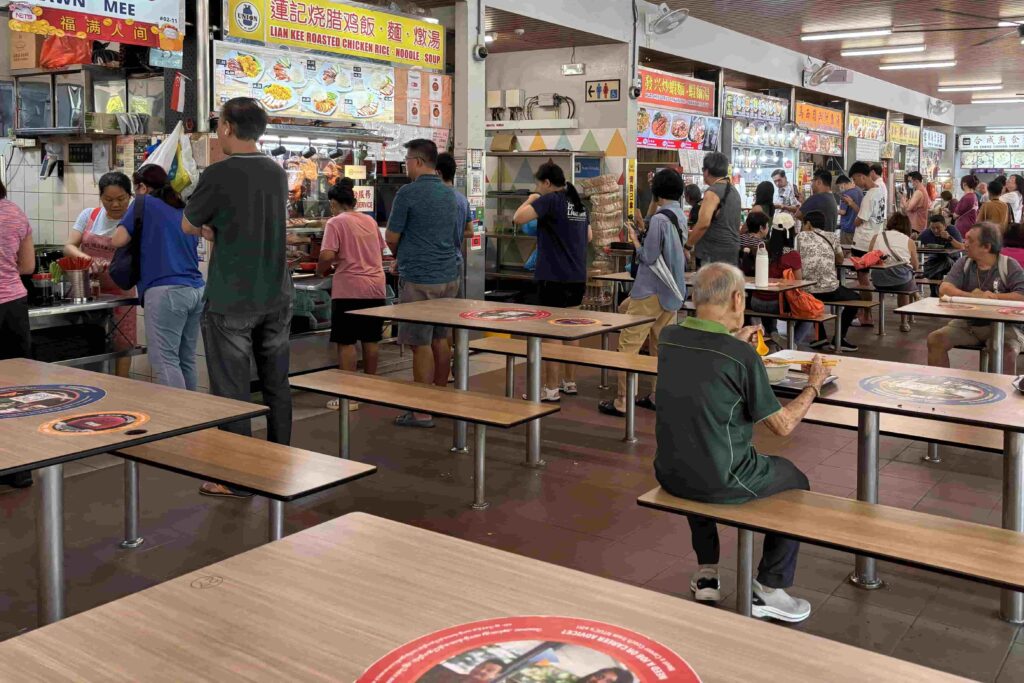 A busy Singapore hawker centre with people queuing at food stalls and diners eating at communal tables.