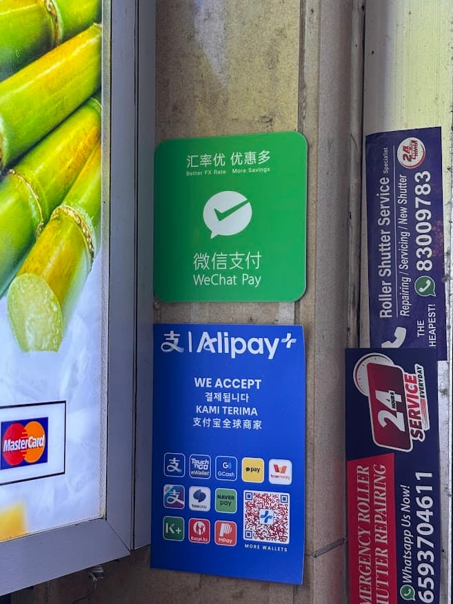 Close-up of digital payment signage at a Singapore hawker stall showing green WeChat Pay and blue Alipay+ decals displayed on a concrete pillar next to a sugarcane drink stall.