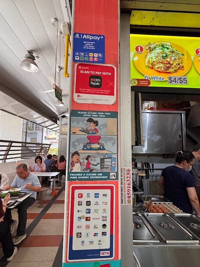 A Singapore hawker centre stall pillar displaying a red SGQR code decal for NETS and local banking apps, alongside a hawker food menu board showing carrot cake pricing, with diners eating in the background.