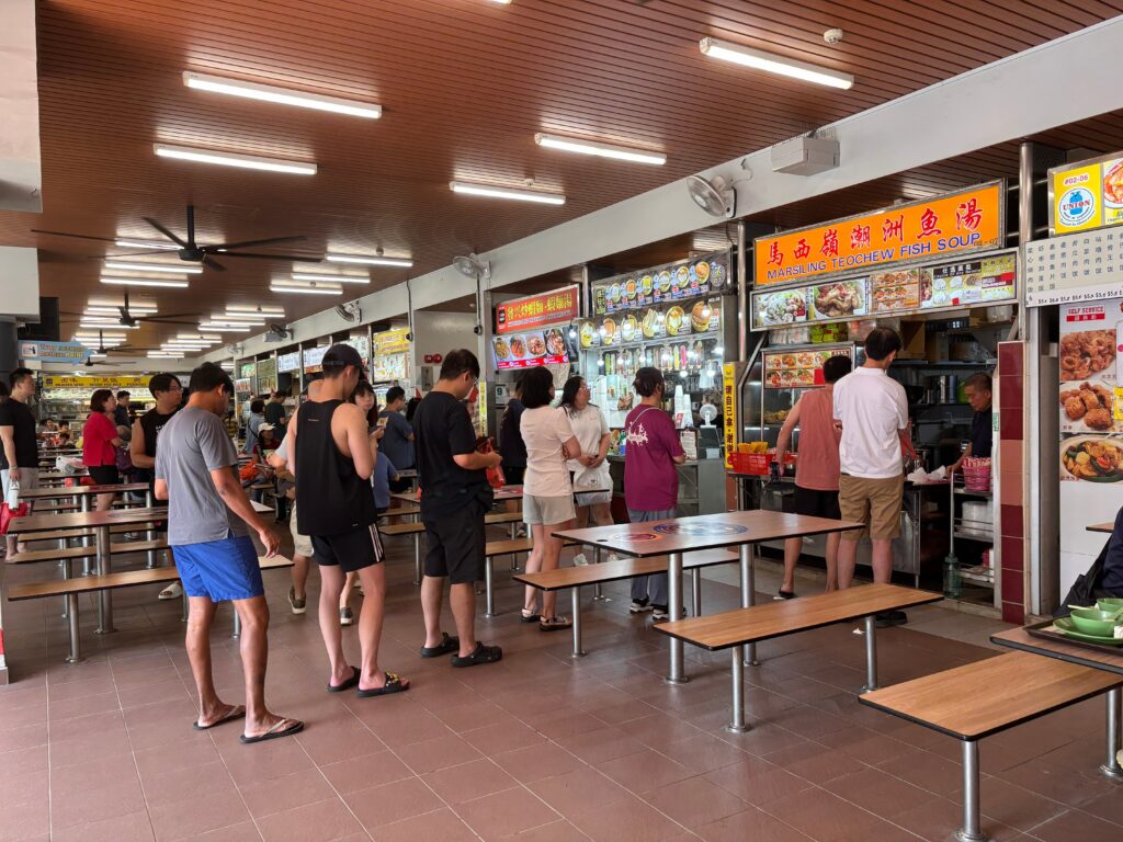 A busy Singapore hawker centre with a queue of people waiting to order from the popular fish soup stall.
