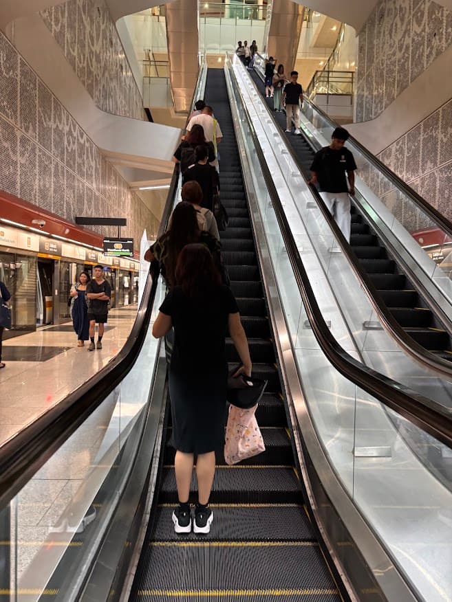 Commuters standing on the left side of a long escalator in a Singapore MRT station, leaving the right side open for people walking up.
