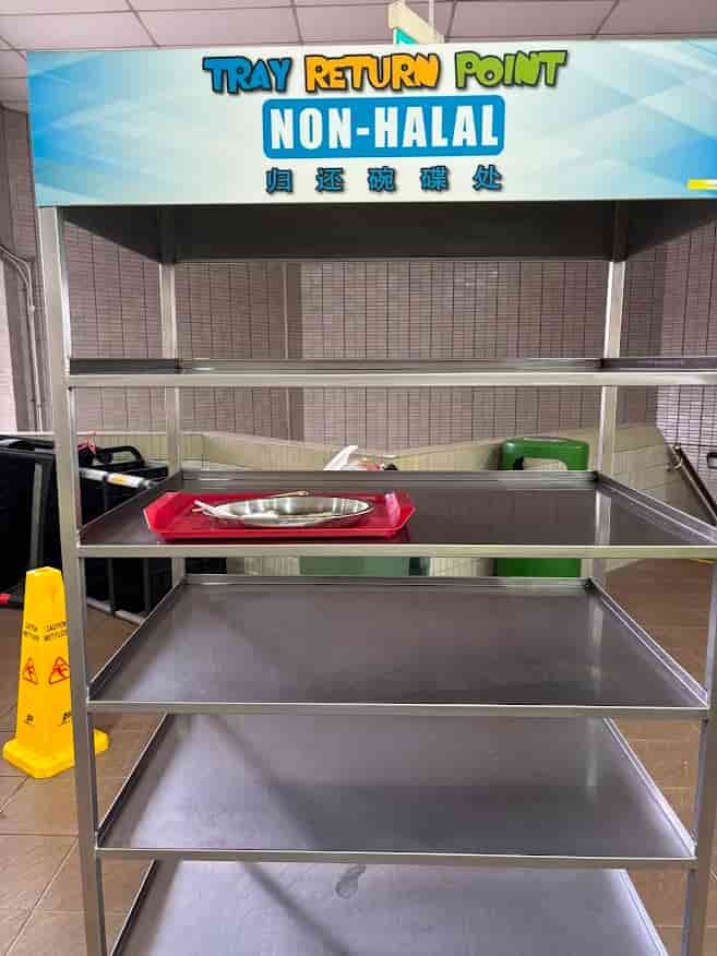 A metal multi-tiered tray return shelf at a Singapore hawker centre with a large blue sign reading "TRAY RETURN POINT" and "NON-HALAL.