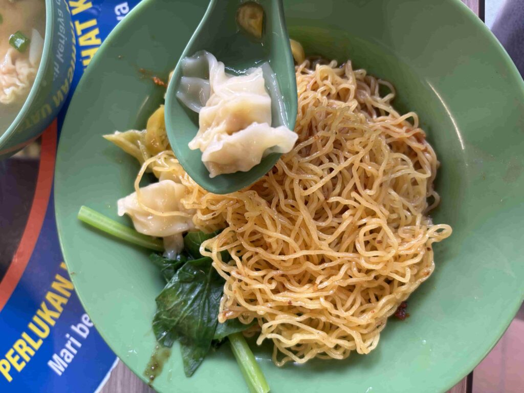 Wonton Mee, a Hawker Favourite. A bowl of Singapore wonton mee noodles with green leafy vegetables and a spoon holding a wanton dumpling.