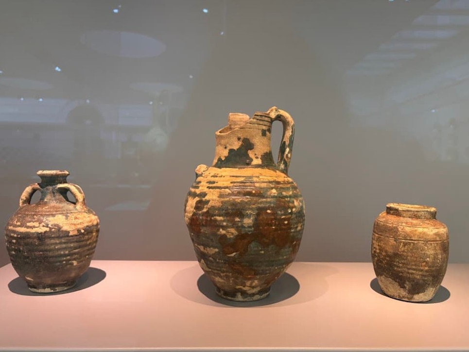 Three earthenware vessels from the Abbasid Empire on museum display — two small jars and a larger amphora with a handle, all showing traces of turquoise glaze and age.
