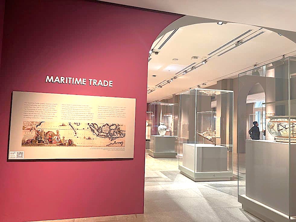 The entrance to the Maritime Trade gallery at the Asian Civilisations Museum, featuring a maroon accent wall with a large informational display board under the text MARITIME TRADE, opening into a bright exhibition room with glass showcases.