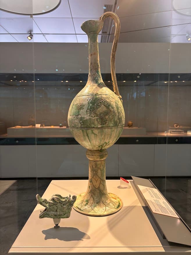 A tall ceramic ewer from Tang Dynasty China, displayed in a museum. The vessel has a long elegant neck, a curved handle, and decorative incised patterns on the body