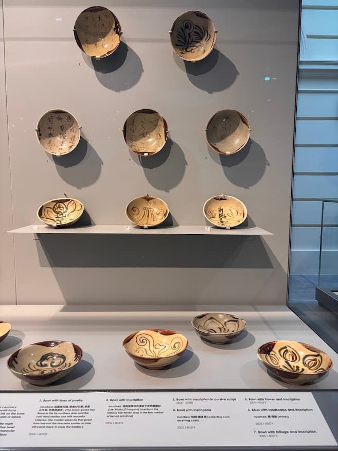 A museum display showing multiple Changsha bowls arranged on a dark wall, with additional bowls and ceramic pieces on a shelf below.