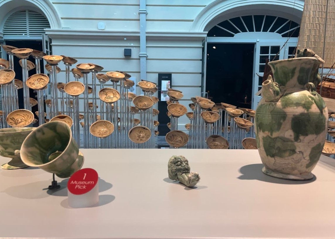 A museum display showing Tang Shipwreck stoneware including a green-glazed pitcher with a feline handle and dragon-head spout, a small animal-shaped incense burner foot, and rows of ceramic bowls mounted on rods in the background.
