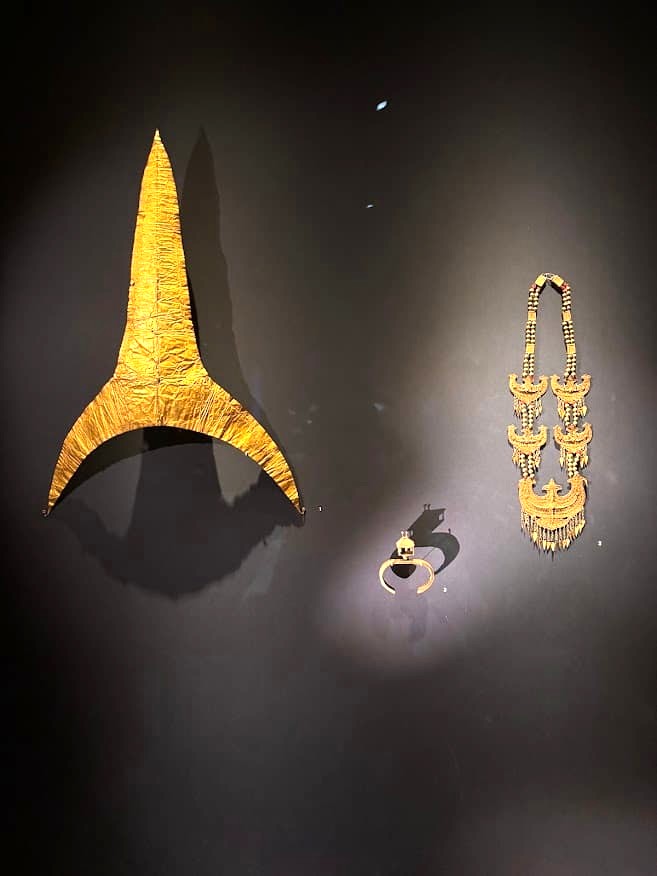 Two gold jewellery pieces displayed against a dark background at the Asian Civilisations Museum Singapore — a pointed gold headdress ornament and an elaborate gold pendant necklace.