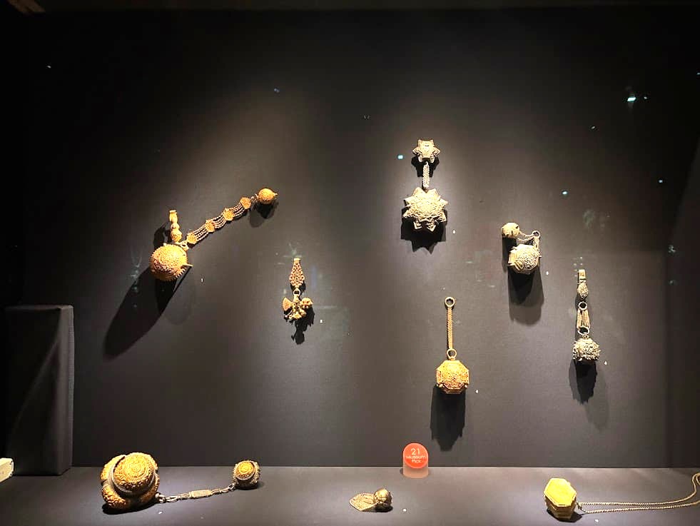 A museum display of small gold and gemstone jewellery pieces including earrings, pendants, and decorative ornaments mounted on a dark wall at the Asian Civilisations Museum Singapore.