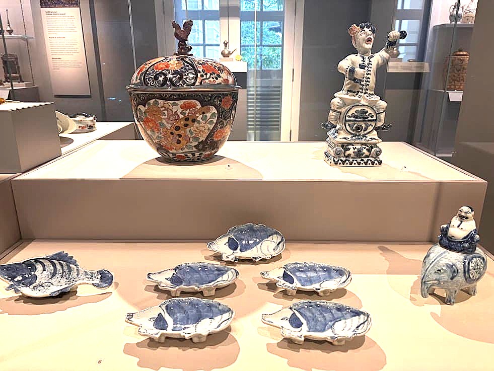 A museum display of blue-and-white ceramic pieces including small boat-shaped dishes, a large decorative vase, and a figurine, arranged on a white display shelf.