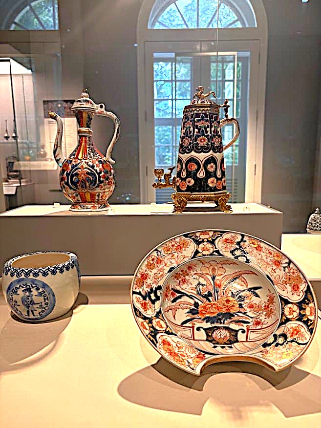 A museum display of Asian export ceramics including an Imari-style pitcher, a large tankard with gold metal mounts, and a decorative plate with blue, red, and gold floral patterns.