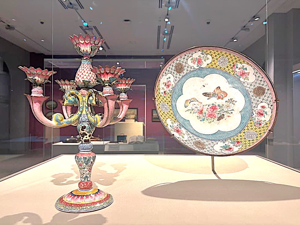 A decorative Chinese candelabra with five lotus-shaped holders and a large enamel dish painted with flowers and butterflies, both displayed in a museum case.