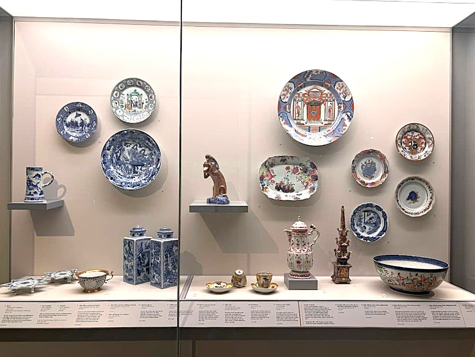 A museum display case showing a collection of Chinese export porcelain including decorated plates, bowls, vases, and small ceramic pieces arranged on shelves and wall mounts.