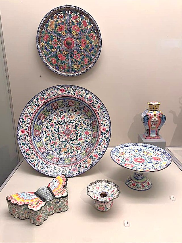 A museum display of 18th-century Guangzhou painted enamel on copper pieces, including a decorative plate, a pedestal dish, a small vase, a butterfly-shaped inkwell, and a bowl.