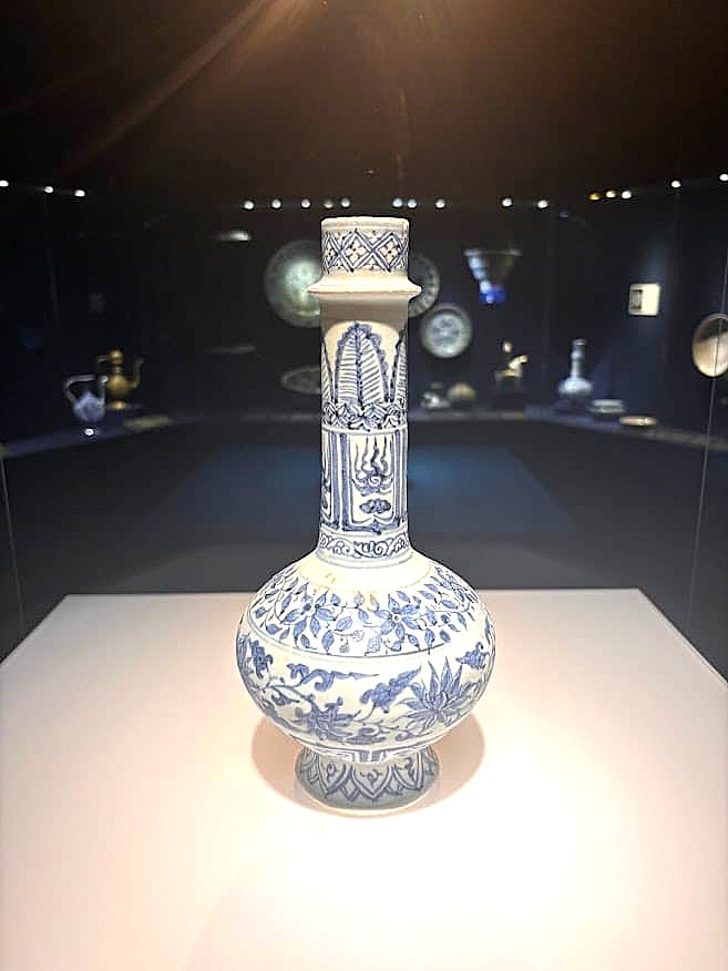 A tall blue-and-white porcelain bottle with a long neck and floral scrollwork decoration, displayed in a dimly lit museum case.