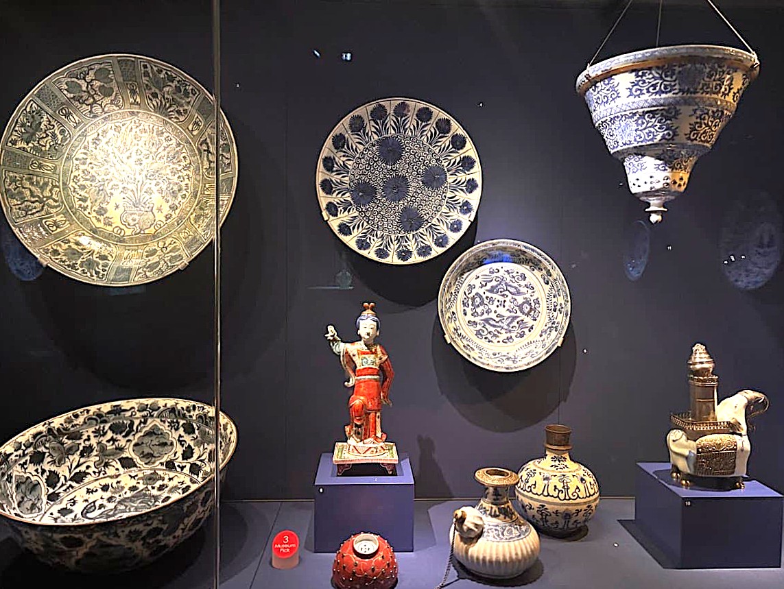 A dimly lit museum display of Chinese porcelain pieces including decorative plates, bowls, and small ceramic objects, some with intricate pierced patterns and gold accents.