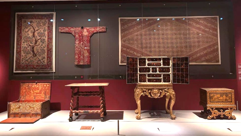 A museum display at the Asian Civilisations Museum Singapore showing antique furniture and textiles including decorative wall panels, a red ceremonial robe, ornate chairs, and carved wooden chests.