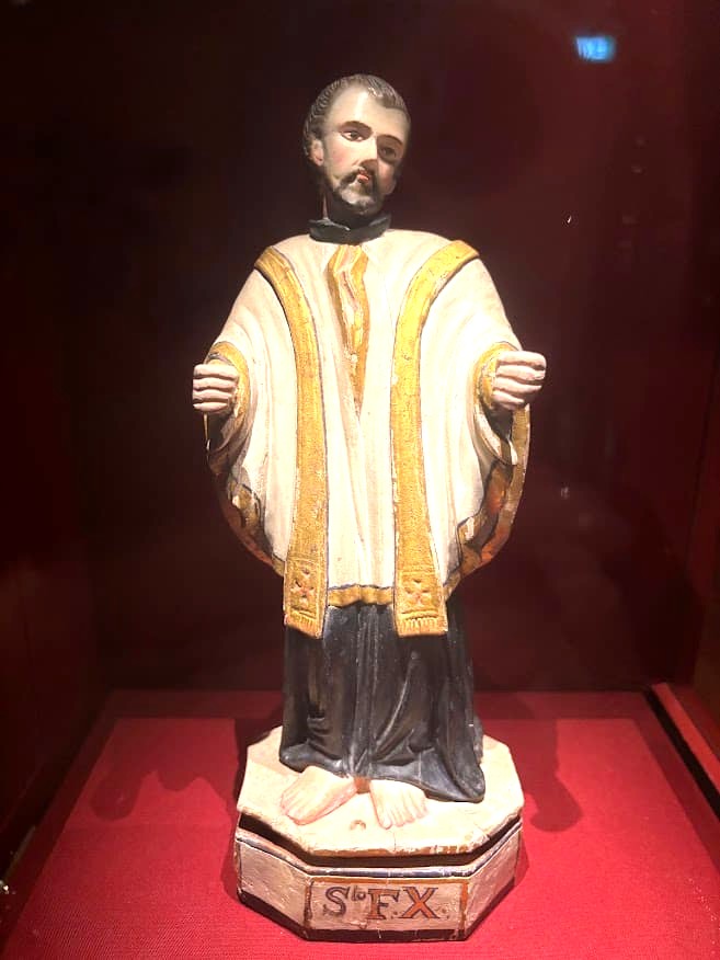 A carved and painted statue of Saint Francis Xavier in white and gold robes on a decorated base inscribed with "S.FX", on display at the Asian Civilisations Museum Singapore.