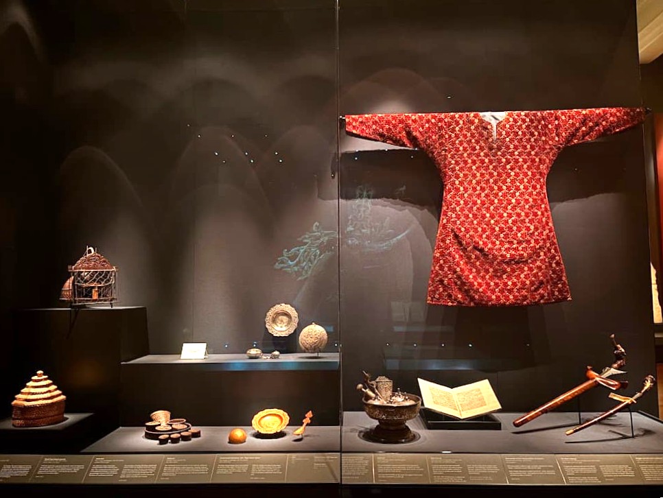 A museum display at the Asian Civilisations Museum Singapore showing Malay cultural objects including a red ceremonial robe, ornate betel nut accessories, a sword, and decorative vessels arranged on a dark display table.