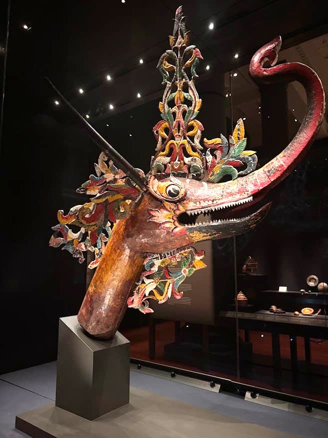 A ornate wooden makara processional vehicle on display at the Asian Civilisations Museum Singapore, depicting a mythical creature with crocodile, fish, goat and elephant features, decorated in vivid colours and gold.