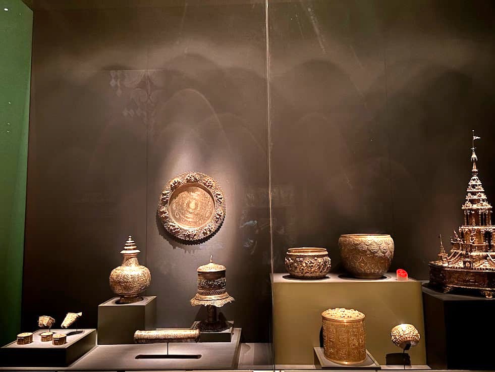 A museum display at the Asian Civilisations Museum Singapore showing a collection of dark bronze or brass vessels, bowls, and ceremonial objects arranged on a shelf.