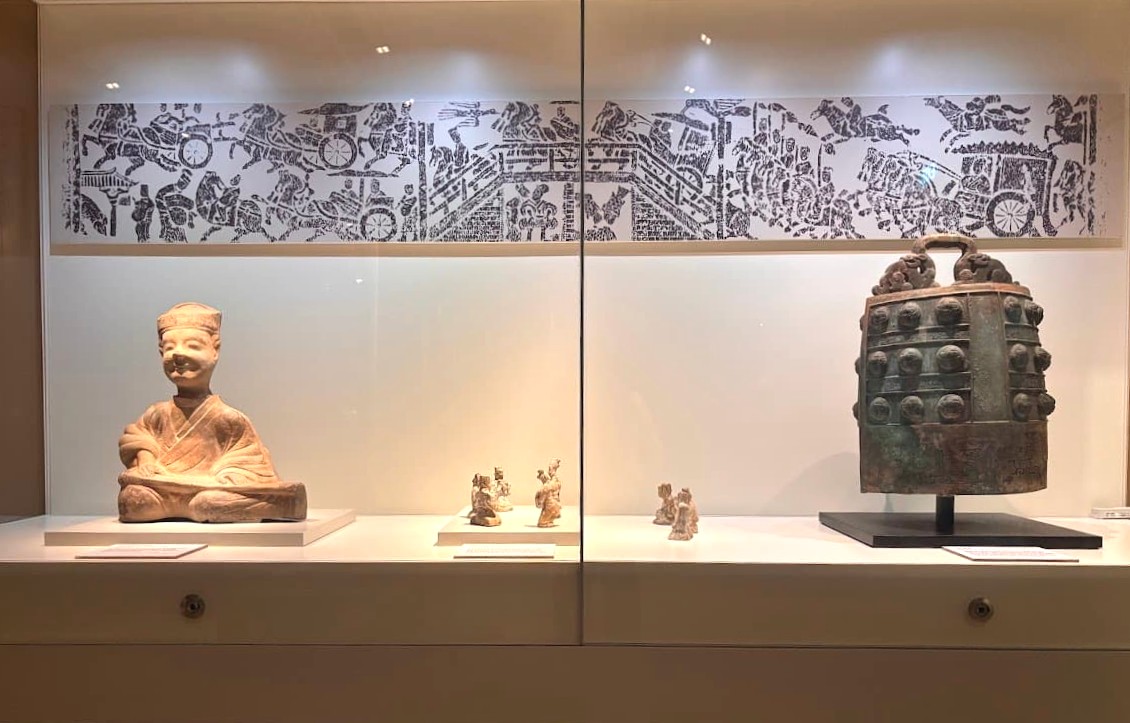 A museum display at the Asian Civilisations Museum Singapore showing a large carved relief panel, small decorative figurines, and a bronze vessel in the Scholars Gallery.