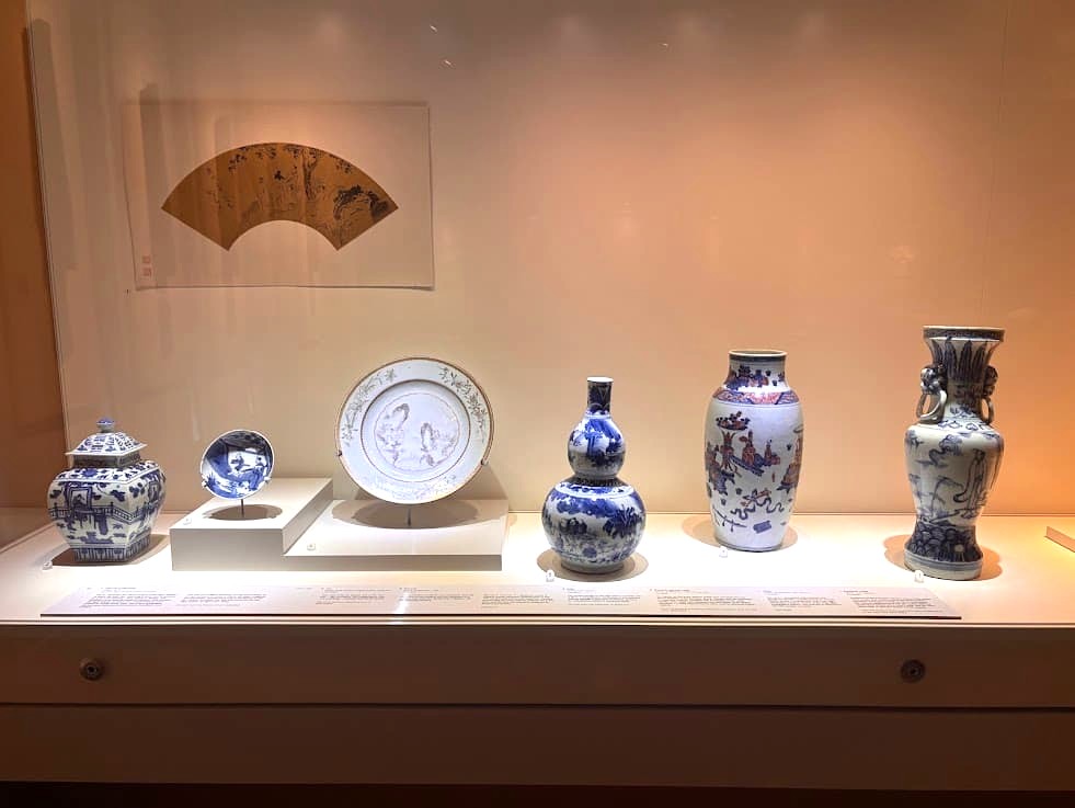 A museum display at the Asian Civilisations Museum Singapore showing a collection of blue-and-white Chinese porcelain including vases, a gourd-shaped bottle, decorative plates, and a lidded jar, with a painted fan artwork mounted on the wall above.