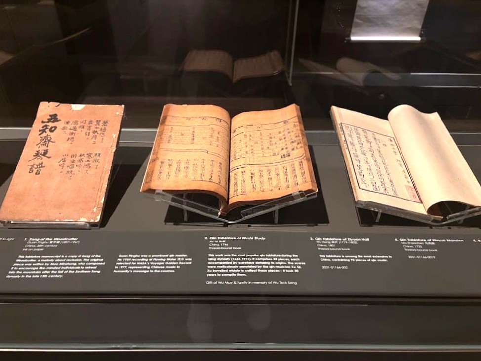 A museum display of four ancient Chinese qin tablature books open on a dark display case at the Asian Civilisations Museum Singapore, showing handwritten musical scores and text.