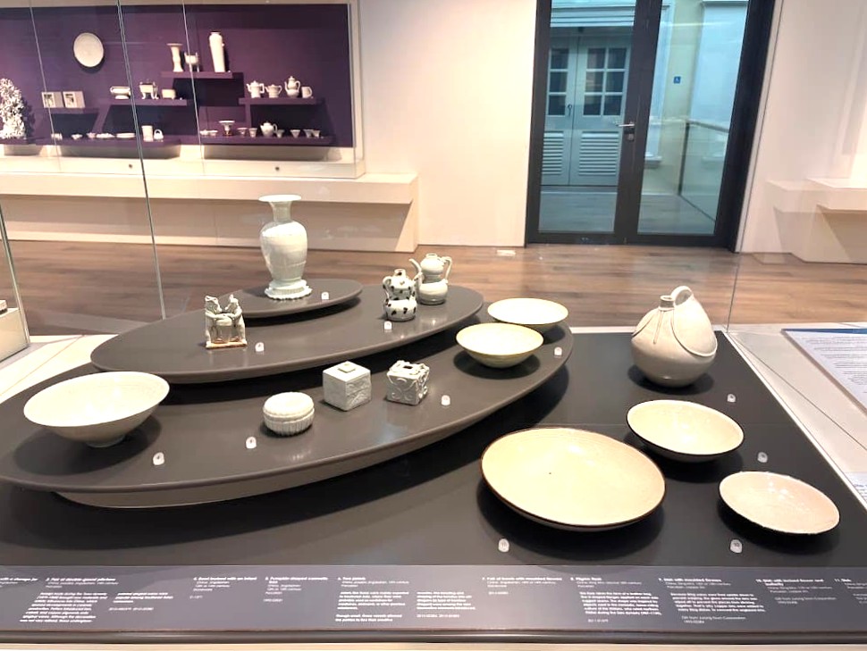 A museum display of Song and Tang dynasty pale celadon and white glazed Chinese ceramics including bowls, vases, pitchers, boxes, and figurines arranged on dark oval display platforms at the Asian Civilisations Museum Singapore.