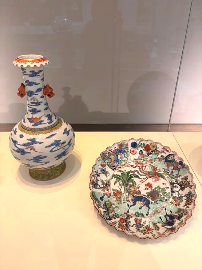 Two Chinese porcelain pieces on display at the Asian Civilisations Museum Singapore — a Qianlong-period vase decorated with bats amid clouds, and a Kangxi-period dish painted with auspicious creatures in vivid wucai colours.